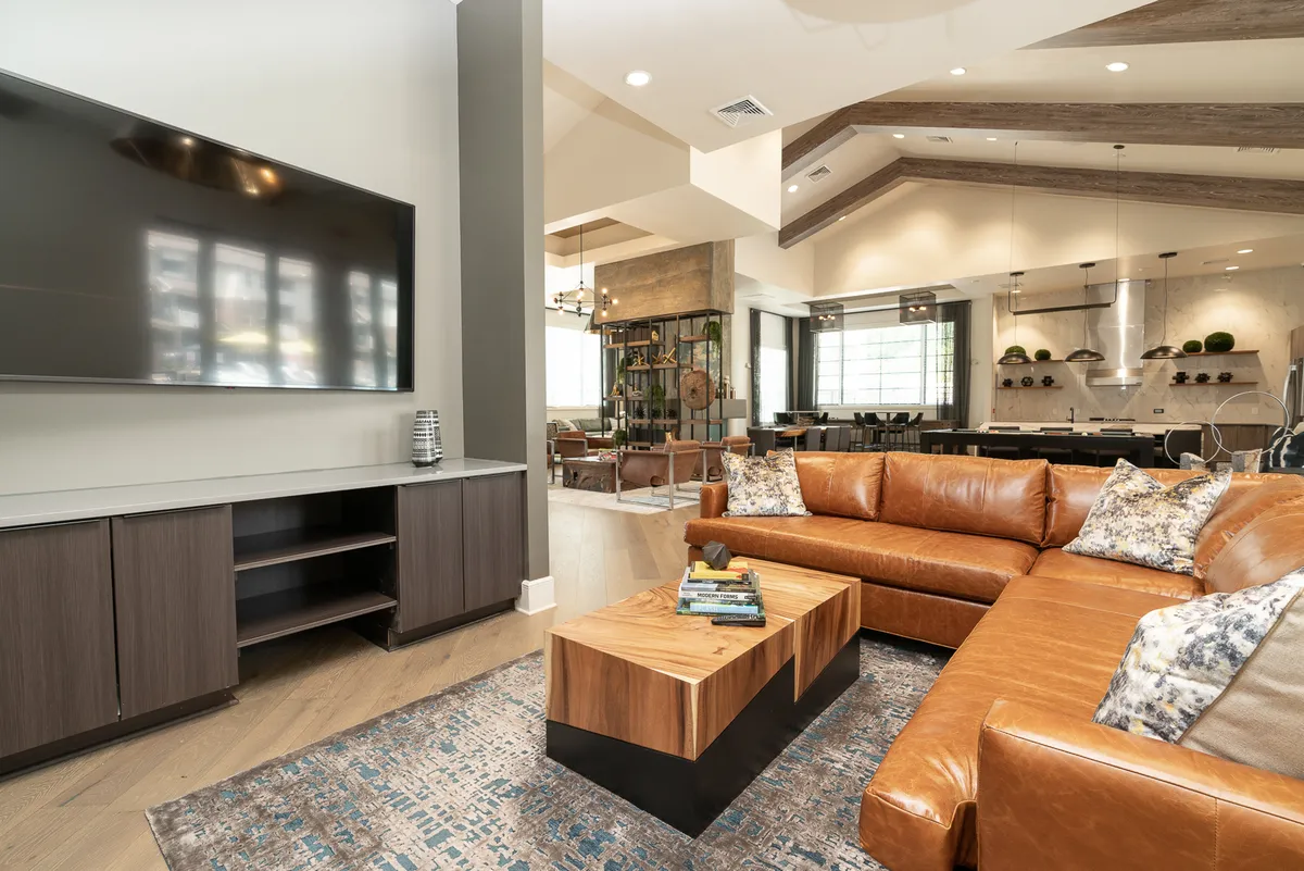 A spacious, open-plan living area featuring a large leather sectional sofa adorned with decorative pillows, a sleek modern coffee table, and a flat-screen TV. In the background, a stylish dining area can be seen, highlighting the apartment's thoughtful design and comfort-focused furnishings.