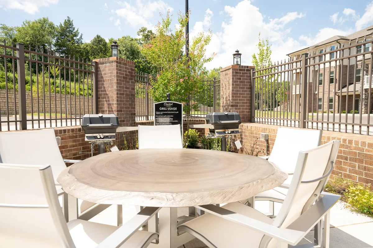 An inviting outdoor dining area featuring a round wooden table, white chairs, grill stations, and lush greenery. The space is designed for al fresco dining or relaxed gatherings, making it ideal for celebrating special occasions or simply unwinding together.