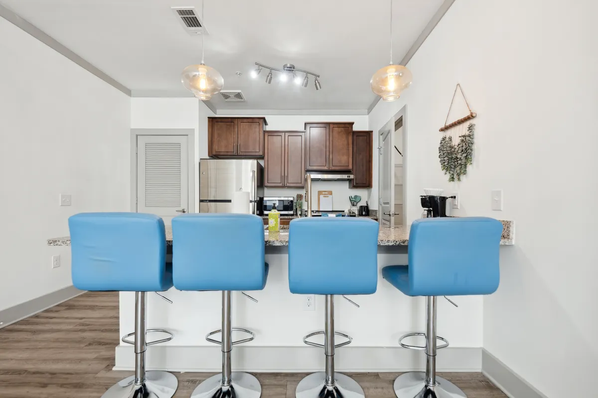 A contemporary kitchen and dining area with blue bar stools along a granite-topped island, complemented by warm wooden cabinetry and state-of-the-art kitchen appliances. The scene is lively yet welcoming, suggesting a space designed for both entertaining and everyday living.