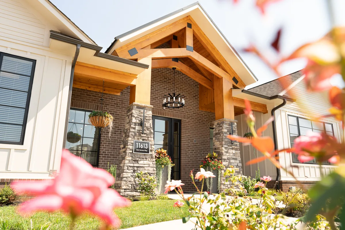 A striking entrance arch framed by stone and adorned with a chandelier, flanked by blooming flower beds and a stone facade. The setting hints at both elegance and warmth, perfect for welcoming guests or stepping out for a city adventure.