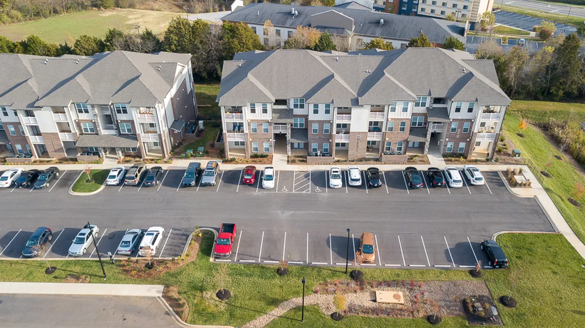 A multi-story apartment building set against a backdrop of autumn foliage, with a landscaped garden area in the foreground. Residents can be seen walking, parking their vehicles, and socializing in the garden, creating an inviting and community-oriented atmosphere.