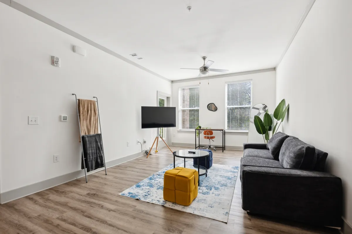 A stylish living room featuring a comfortable sofa, modern coffee table, television, desk, and a lush plant. The space is bathed in natural light and designed for relaxation, working, or entertaining guests. The arrangement emphasizes an open-concept feel, blending lounge and work areas seamlessly.