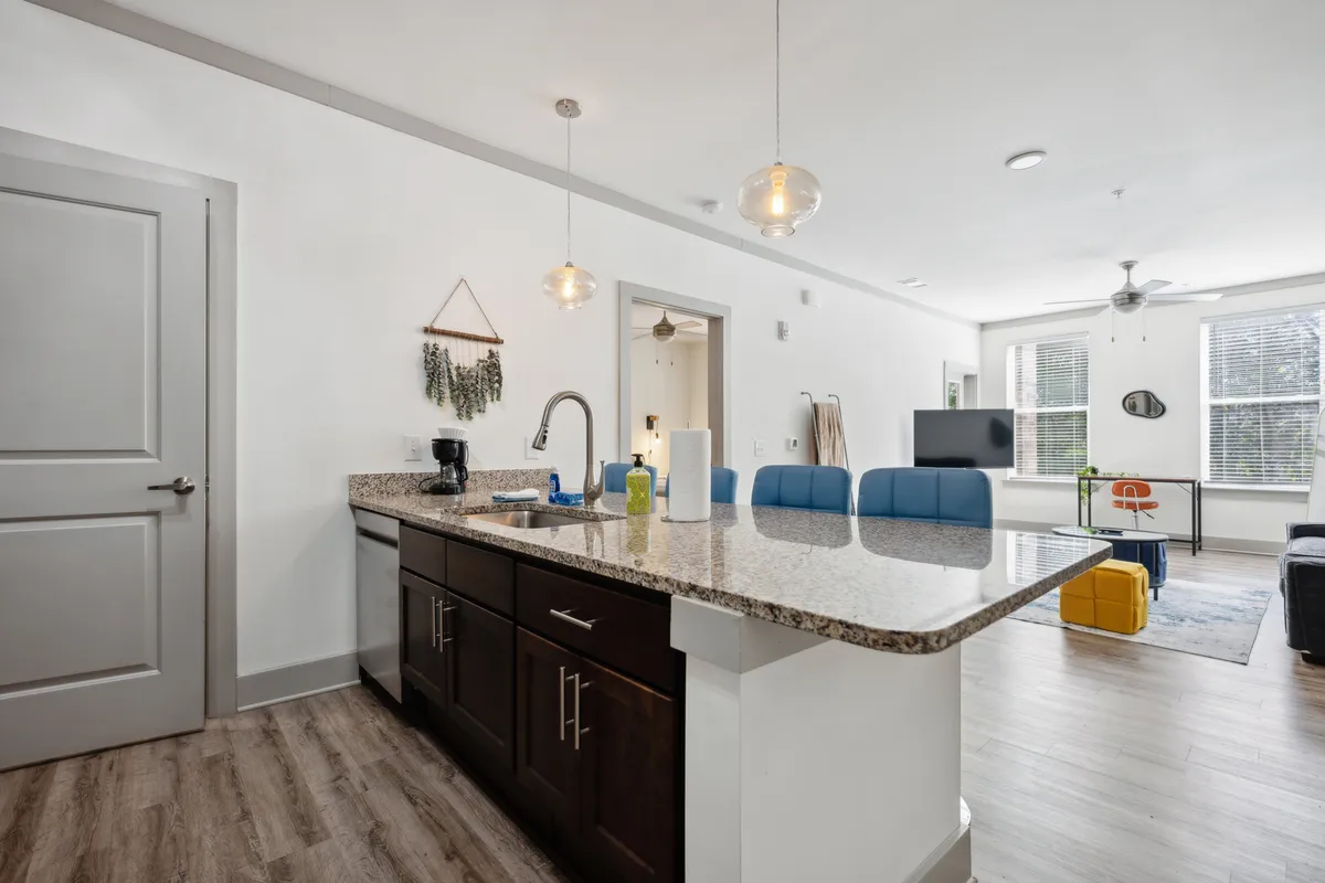 The image showcases a bright, modern apartment interior featuring a sleek granite countertop accented by stylish blue bar stools. The open-plan layout connects the contemporary kitchen space seamlessly to a cozy living area, suggesting an inviting space for cooking, casual dining, and relaxing.