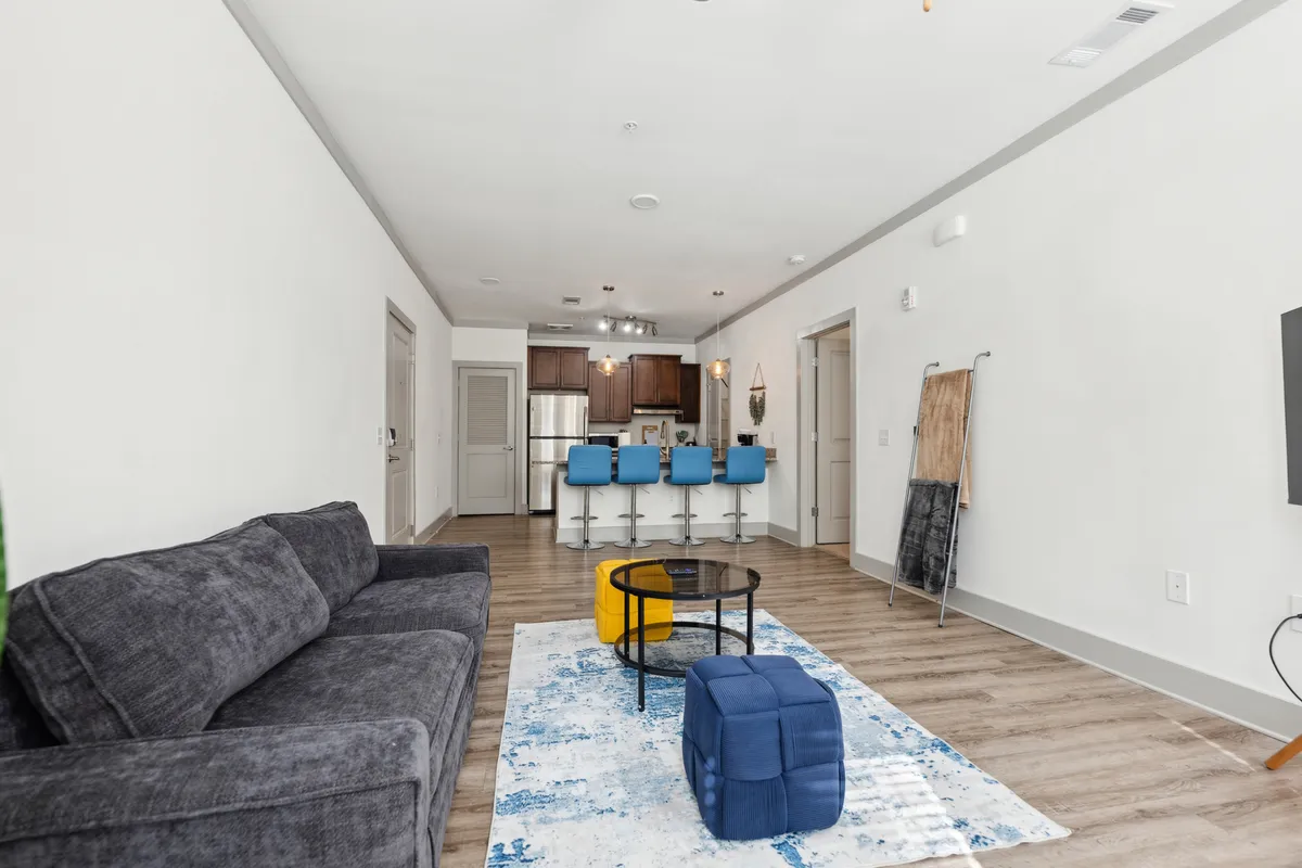 A modern, open-plan living space with a gray sofa, blue accent stools, round coffee table, playful poufs, and a view into the kitchen. The space is lively and uncluttered, perfect for both relaxing and hosting guests.