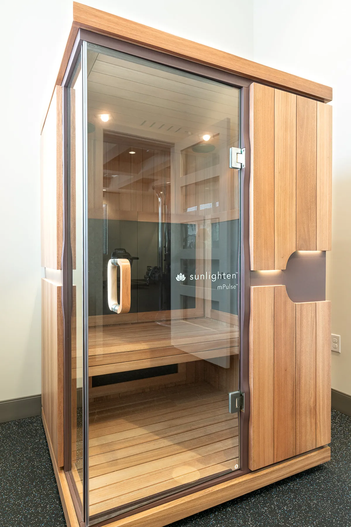 This image showcases a modern infrared sauna set within sleek, smooth wooden panels and a glass door, exuding a tranquil vibe. The soft lighting and clean lines create a sense of wellness and quiet escape, inviting viewers to imagine a soothing self-care session.