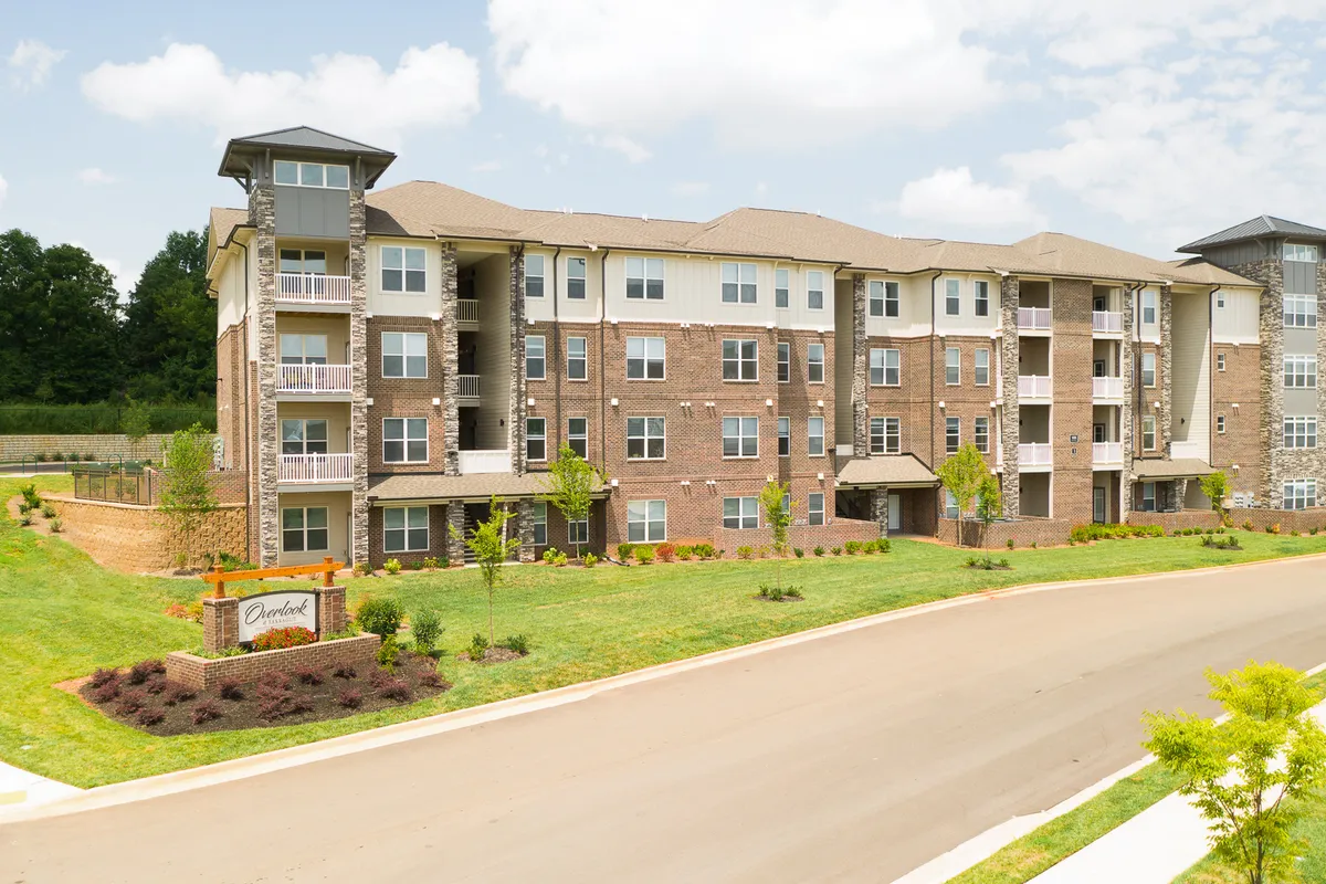 A modern apartment building surrounded by manicured gardens and an entrance sign, with sunny lawns and pathways that encourage outdoor leisure and evening strolls.