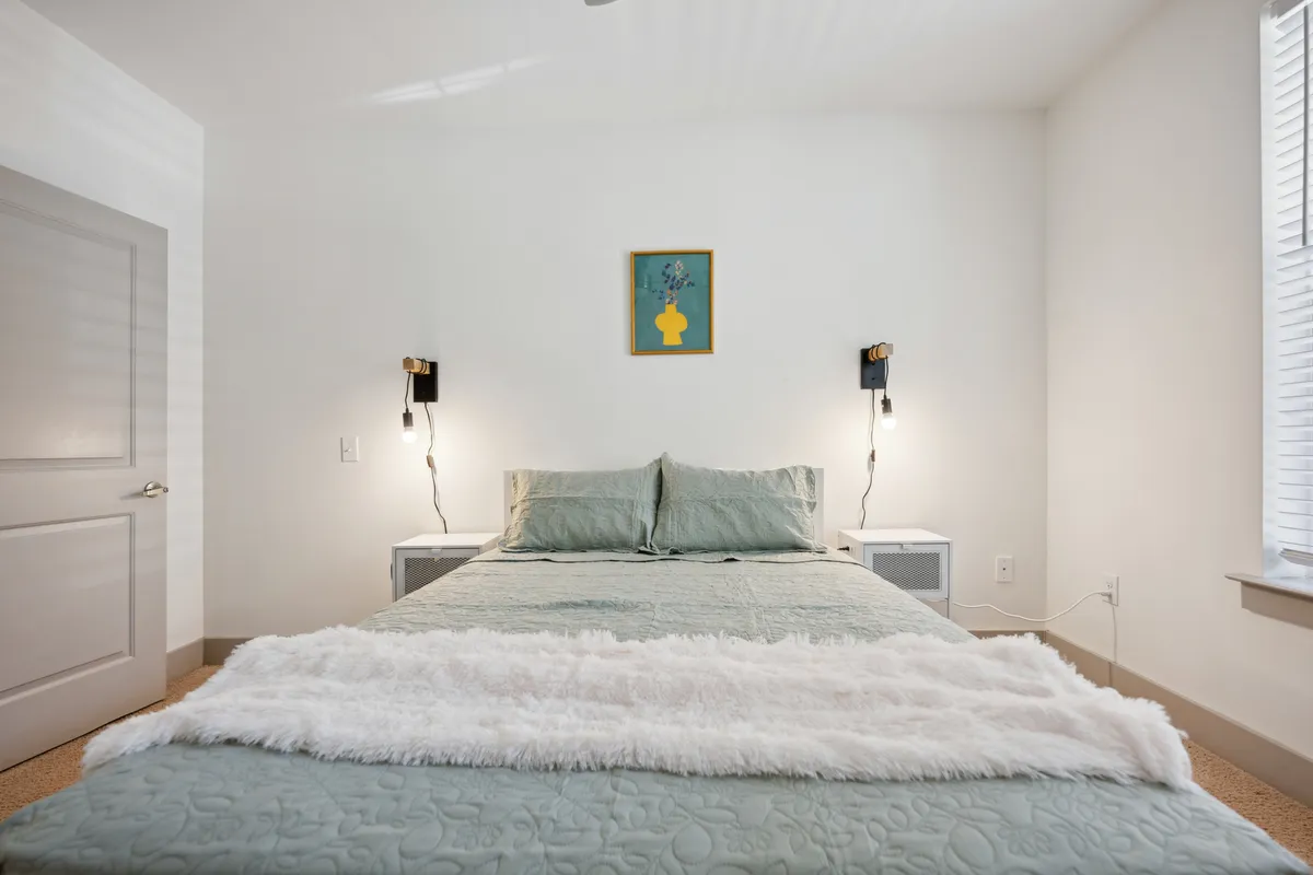A cozy bedroom setting with a bed dressed in light green bedding, two wall-mounted lamps for soft illumination, and framed artwork above the headboard. Morning light streams in, creating a warm, inviting atmosphere.